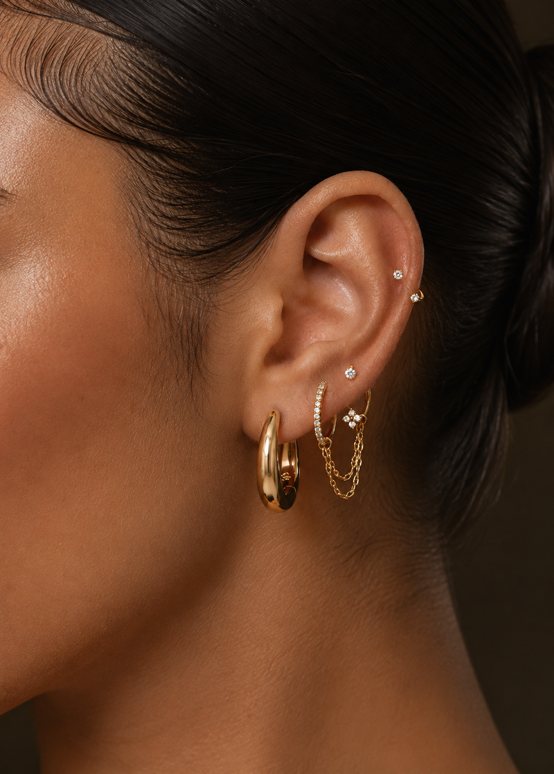 Oreille ornée de créoles dorées, de chaînes au lobe et de deux petites pierres à l’hélix.